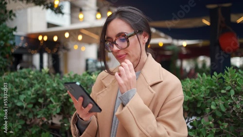 Woman use smartphone outdoor. Portrait caucasian female in glasses using smartphone lloking device screen standing outside dressed cream coat autumn day. People use technology outside, slow motion photo