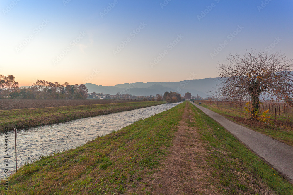 River near a road with colorful trees at sunset in autumn with mountain background, Vittorio Veneto, Italy