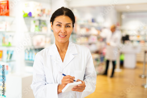 Positive female pharmacist writes in a notebook in the trading floor of the pharmacy