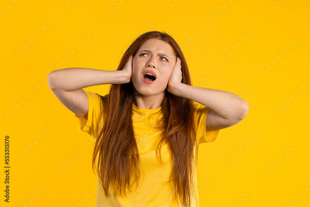 Annoyed ginger woman screaming with shut ears on yellow background. Lady block annoying sounds, music, shouts or noise. Rolling eyes.