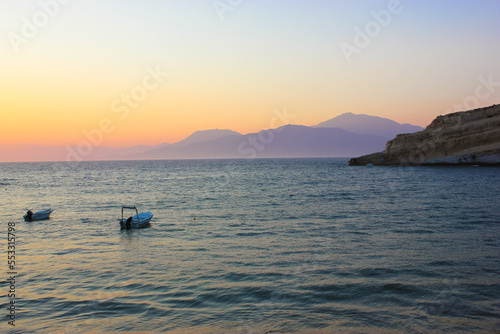 Seascape with silhouettes of hills and mountain. Warm sunlight breaking from behind the mountains at sunrise or sunset. Calm water with small boats. A coast, bay. Serenity, harmony of nature concept.