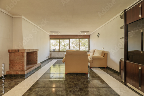 A living room with two-tone stoneware floors, old furniture, a large window and a brick fireplace © Toyakisfoto.photos