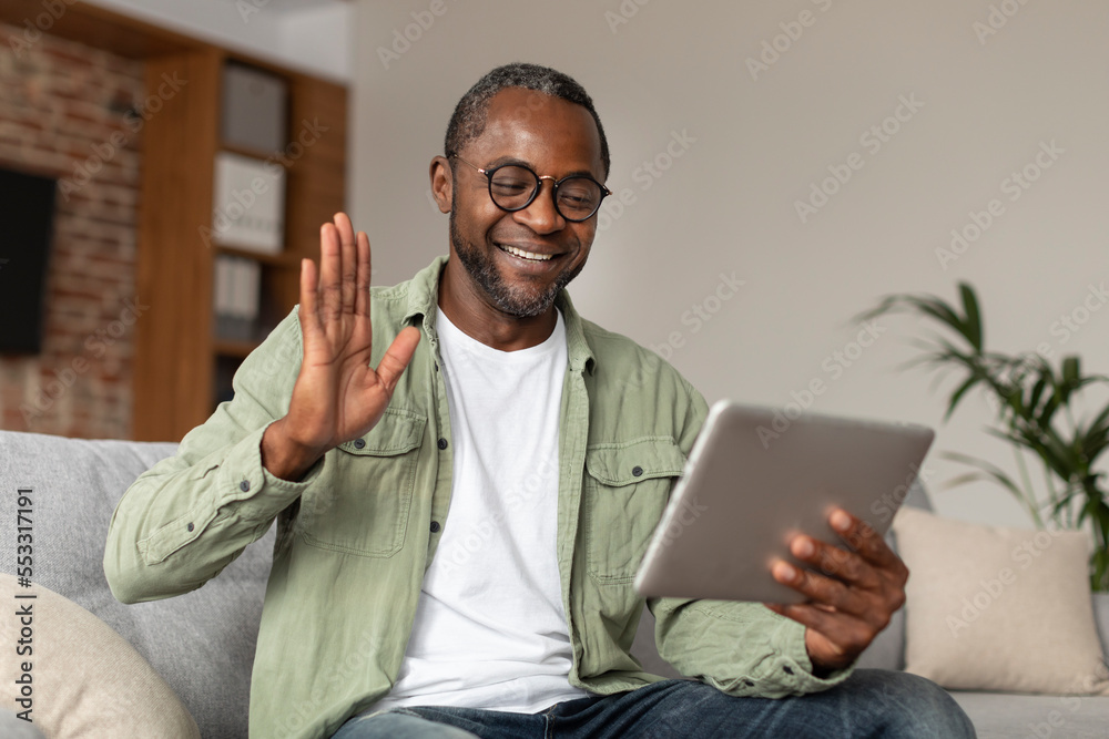 Glad happy middle aged black guy in glasses waving hand at digital tablet webcam, say hello, hi, make greeting gesture - obrazy, fototapety, plakaty 