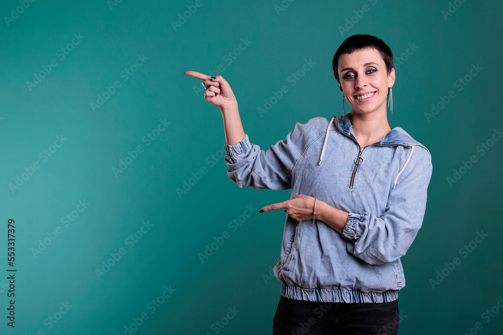 Smiling brunette woman with short hair pointing to side with forefinger advertising product while standing over isolated background. Female with positive facial expression showing promotion Smiling brunette woman with short hair pointing to side with forefinger advertising product while standing over isolated background. Female with positive facial expression showing promotion