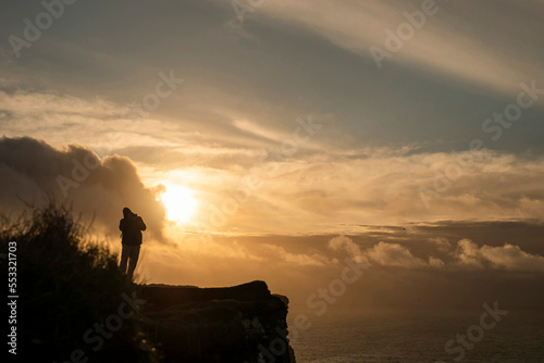 Silhouette of a person standing on a top of a cliff at sunset. Cloudy sky over the ocean. Calm and peaceful mood. Travel and sight seeing.