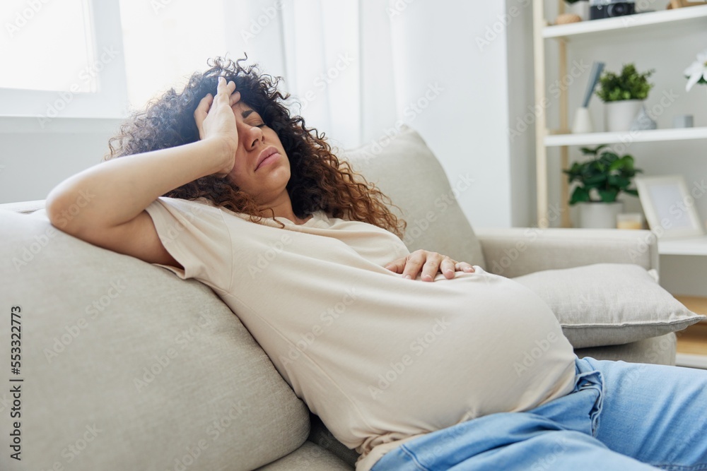 Pregnant woman sitting on the couch at home headache, pregnancy and motherhood difficulties, severe fatigue, headache