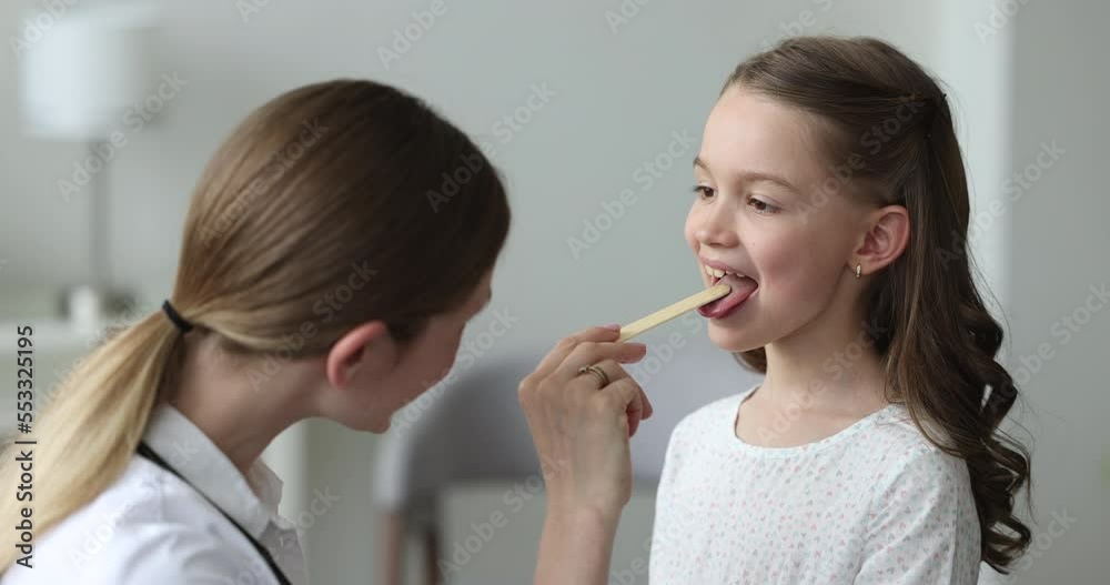 Close up young female ENT otorhinolaryngologist doctor in white coat holding tongue depressor examining little girl throat during visit in private clinic. Medicine, childcare, professional treatment Close up young female ENT otorhinolaryngologist doctor in white coat holding tongue depressor examining little girl throat during visit in private clinic. Medicine, childcare, professional treatment