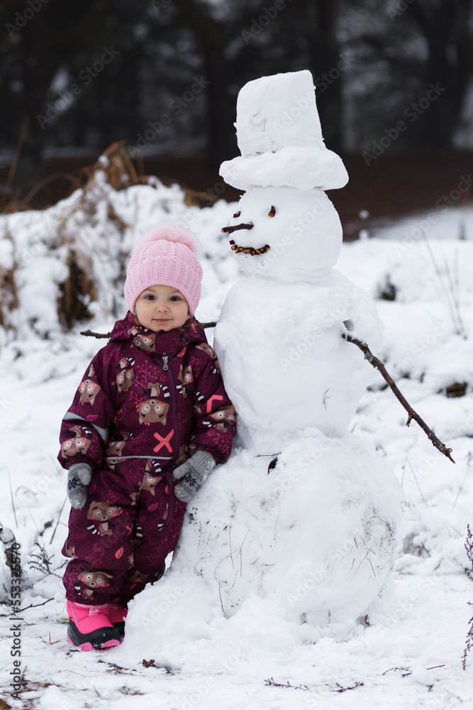 A four-year-old girl stands next to a snowman, there is snow and forest around. Winter, new year.