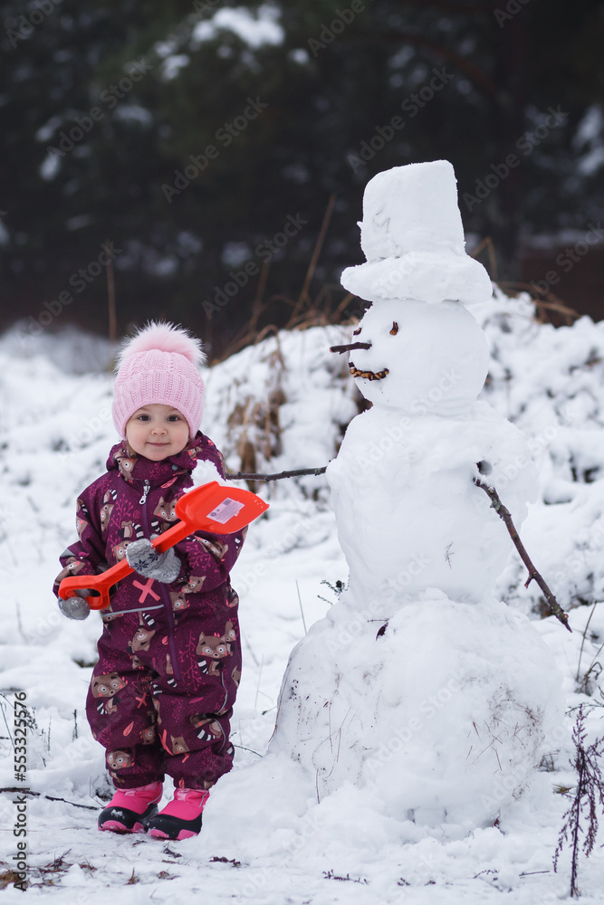 A four-year-old girl builds a snowman in the winter in the forest, holding a red shovel in her hand, surrounded by snowdrifts. Vertical orientation.