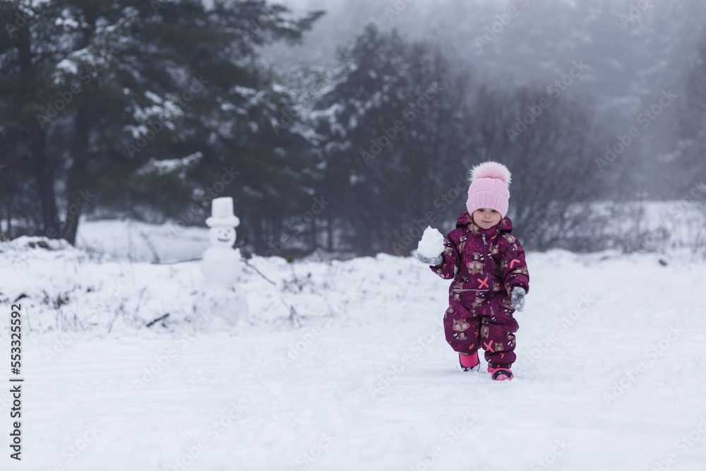 A little girl walks through the snowdrifts in the forest against the backdrop of a snowman. A little girl walks through the snowdrifts in the forest against the backdrop of a snowman.
