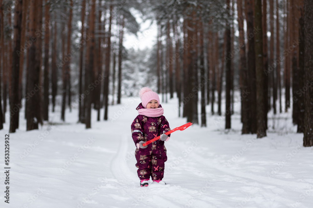 A little girl stands in the middle of a snowy forest with a red shovel in her hands. Looks into the frame. Winter. Snowdrifts are white.