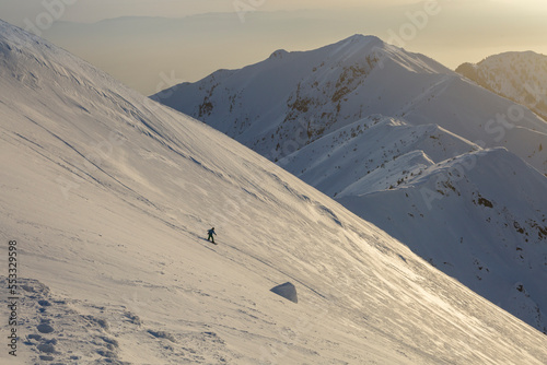 A freerider snowboarder descends a wide slope against the background of mountain ranges and the sunset