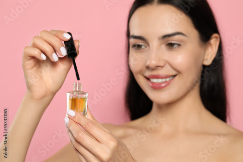 Beautiful young woman with eyelash oil against pink background, focus on bottle