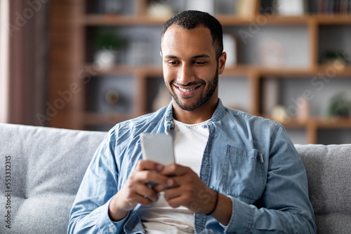 Cheerful Black Man Playing Online Games On Smartphone While Relaxing On Couch,