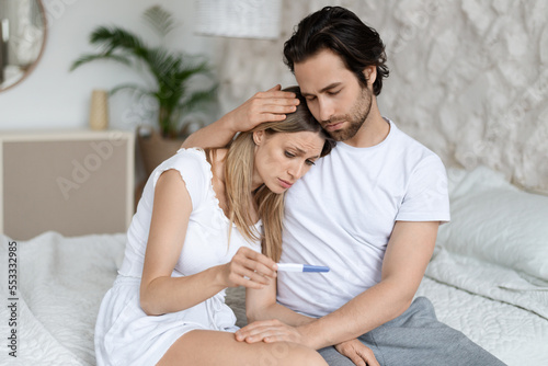 Infertility problem. Caucasian couple sitting on bed with negative pregnancy test result