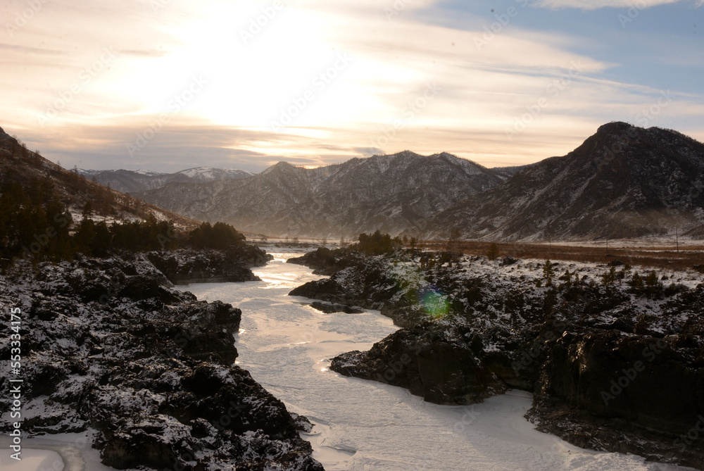 The frozen bed of a narrow river sandwiched in a stone canyon flowing through a snow-covered valley in the mountains on a winter evening before sunset.