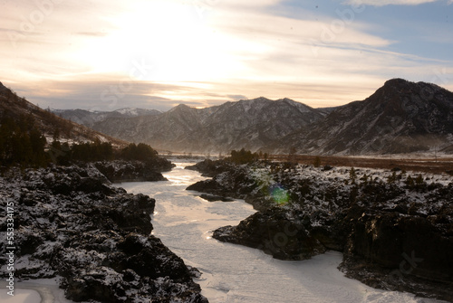 The frozen bed of a narrow river sandwiched in a stone canyon flowing through a snow-covered valley in the mountains on a winter evening before sunset.