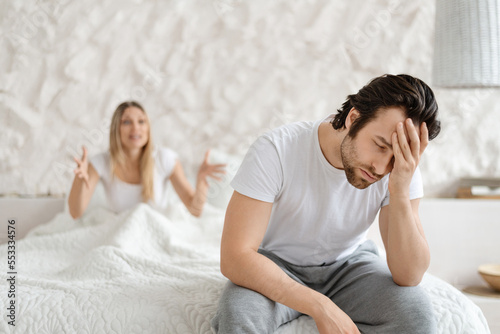 Angry woman shouting at her husband, having conflict while sitting on bed at home, focus on upset man photo