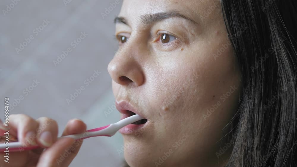 Dark-haired caucasian woman brushing her teeth while standing in the bathroom. Morning ritual concept. Dark-haired caucasian woman brushing her teeth while standing in the bathroom. Morning ritual concept.