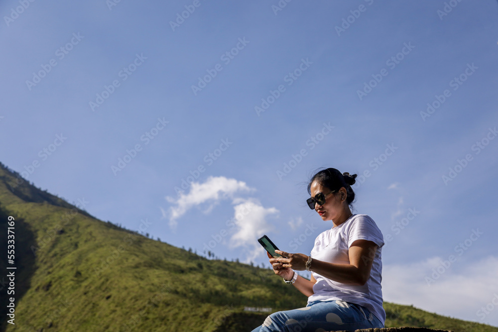 beautiful asian girl in sunglasses sending message on smartphone against green valley and blue sky in bromo plateau background