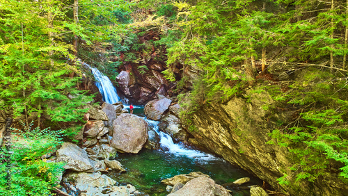 Aerial over river in rocky gorge of lush green forest with waterfall and lone hiker observing falls from boulder photo
