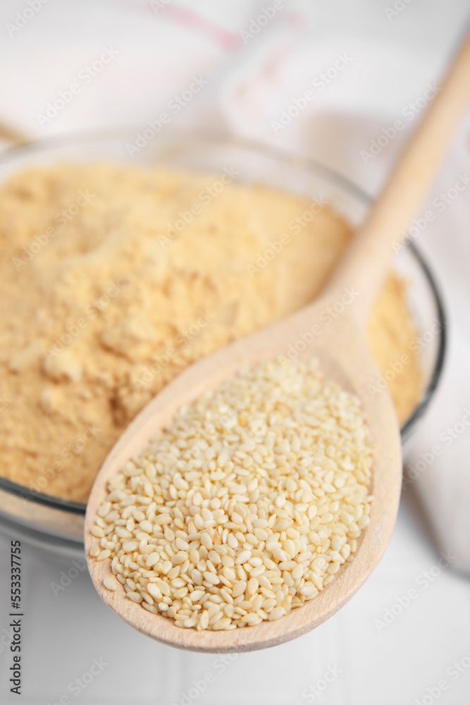 Sesame flour in glass bowl with wooden spoon of seeds on white table, closeup Sesame flour in glass bowl with wooden spoon of seeds on white table, closeup