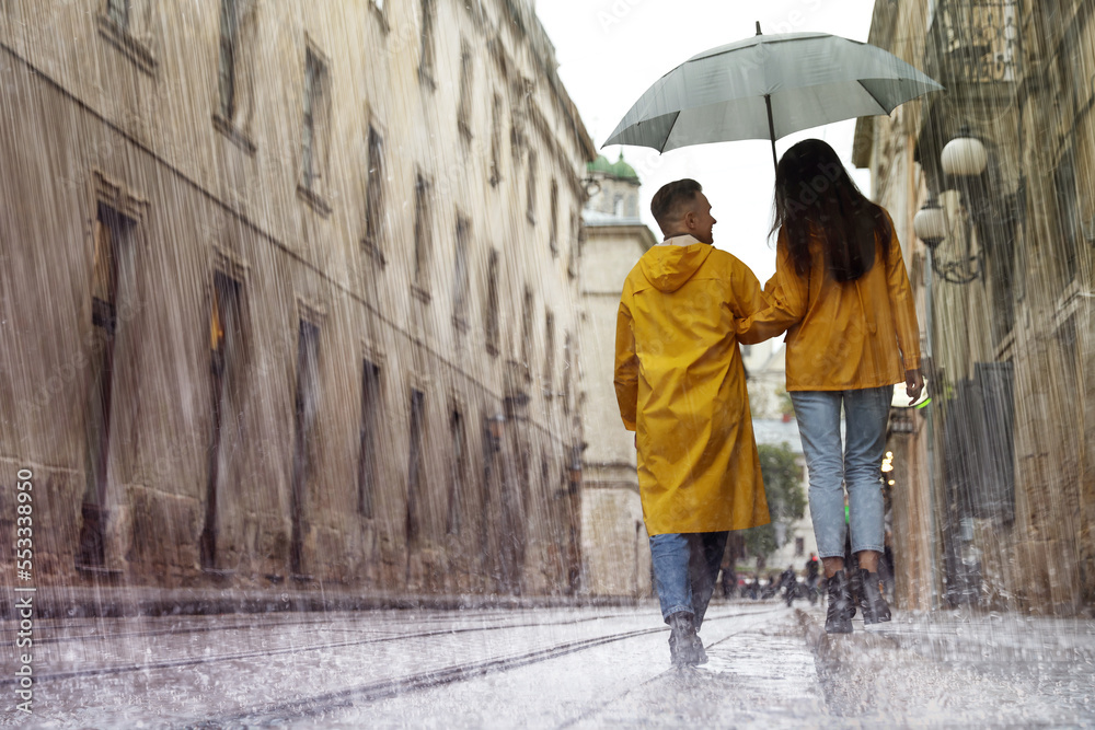 Lovely young couple with umbrella walking under rain on city street, back view