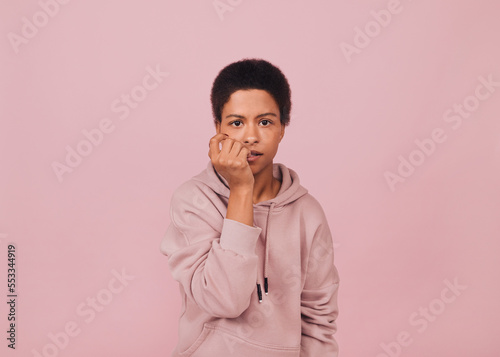 Unsure confused black girl biting her nails. Young woman in casual outfit with pensive face expression against pink background