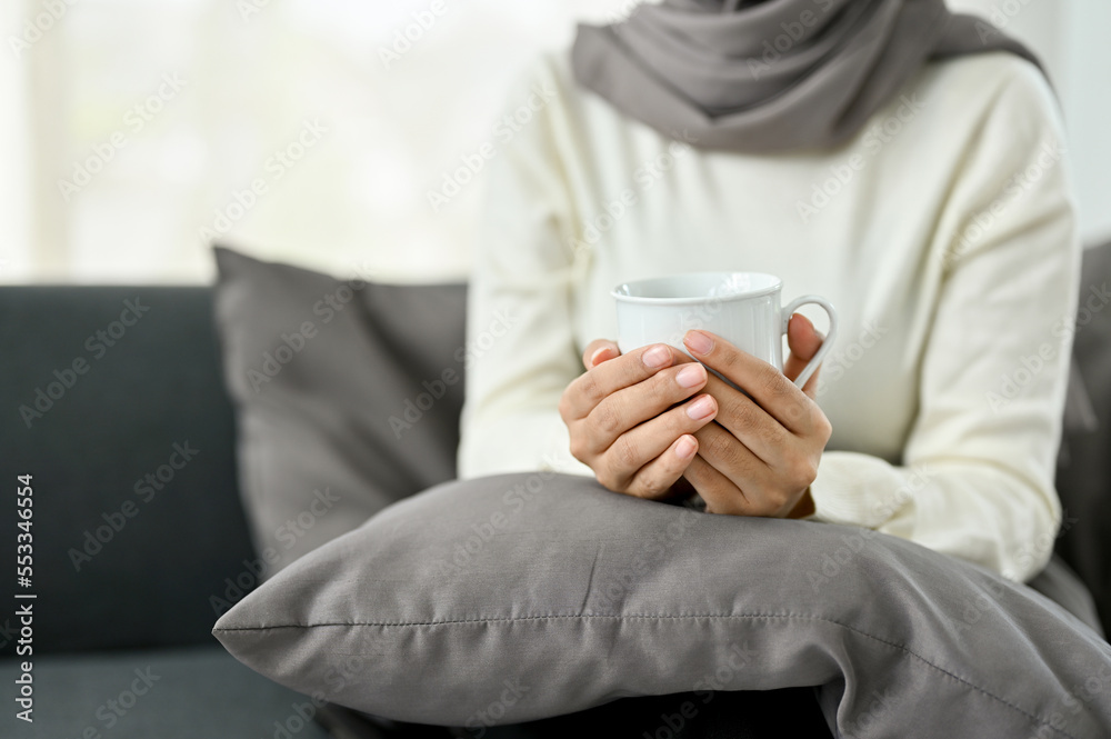 A Muslim woman wearing hijab holding a coffee mug, sitting on sofa in her living room, cropped A Muslim woman wearing hijab holding a coffee mug, sitting on sofa in her living room, cropped