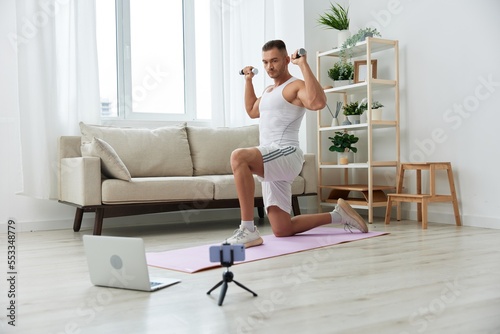 Man sports, watching a workout tape on his phone and repeating exercises sports blogger with dumbbells, pumped up man fitness trainer works out at home, the concept of health and body beauty © SHOTPRIME STUDIO