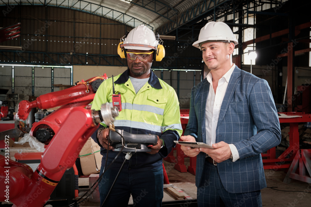 An African American engineer demonstrates smart robotic arm control to customer in mechanical manufacturing factory, intelligent automatic machines in industrial business, and futuristic technology. An African American engineer demonstrates smart robotic arm control to customer in mechanical manufacturing factory, intelligent automatic machines in industrial business, and futuristic technology.