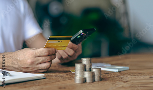 Close-up shot of Asian male hands holding bank card and smartphone. Use an app to shop online. Transfer money and pay with stacks of coins on the table. © ArLawKa
