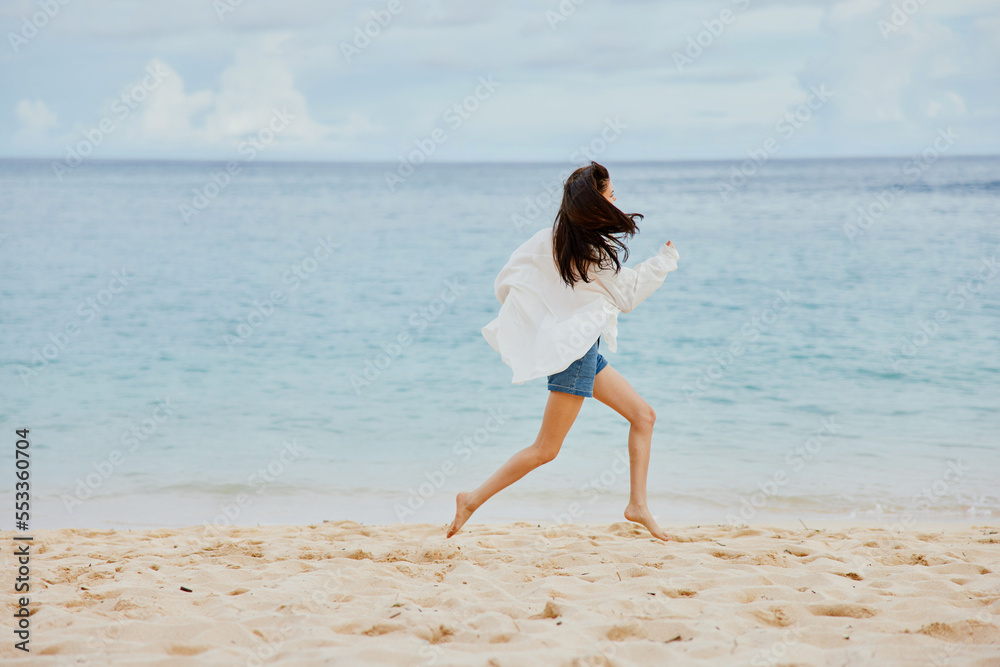 Sports woman runs along the beach in summer clothes on the sand in a yellow T-shirt and denim shorts white shirt flying hair ocean view Sports woman runs along the beach in summer clothes on the sand in a yellow T-shirt and denim shorts white shirt flying hair ocean view