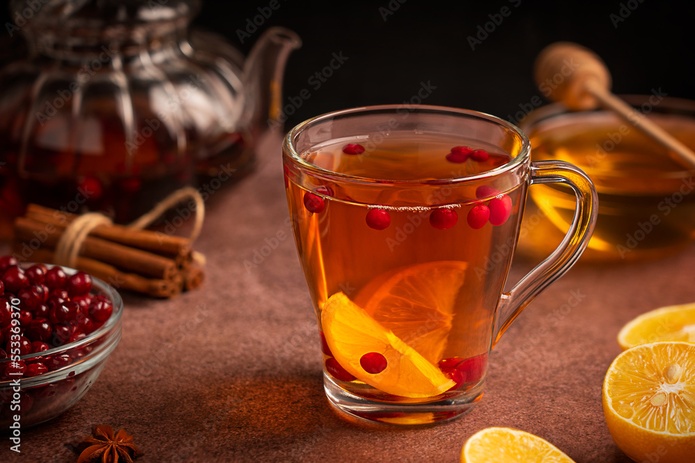 Homemade healthy sweet hot tea drink served in transparent glass cup with red ripe cranberries and lemon citrus fruit slices on dark brown table with teapot, cinnamon spice and honey ingredients Homemade healthy sweet hot tea drink served in transparent glass cup with red ripe cranberries and lemon citrus fruit slices on dark brown table with teapot, cinnamon spice and honey ingredients