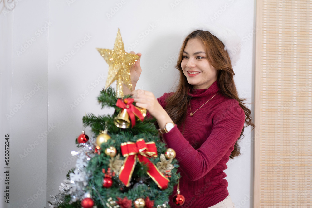 Happy young woman wearing a Santa Claus hat and sweater, smile and enjoy decorating various baubles on the Christmas tree in the living room at home. Enjoying and celebrating the Christmas holiday.