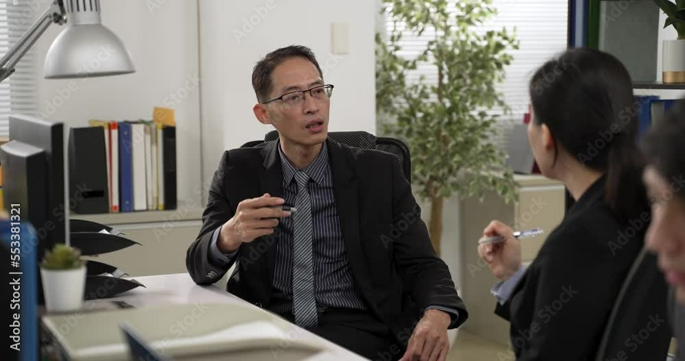 Asian male manger consulting female colleague face to face talking at work in the office with other workers. he uses hand gestures with pen in hand while talking Asian male manger consulting female colleague face to face talking at work in the office with other workers. he uses hand gestures with pen in hand while talking