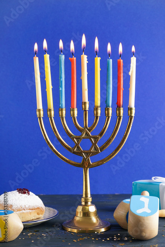 Menorah with doughnut, gift and dreidels for Hannukah celebration on table near blue wall