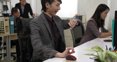 slow motion of young bored asian man employee having energy again when he checks his watch and finds itâs off duty time. he shuts down computer and leaves office happily photo