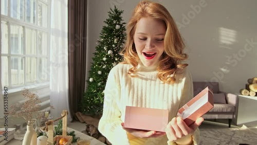 Happy Amazed Woman in Santa Hat, Holding Present in Her Hands, Opens It and Smiles. Next to Xmas Tree. High quality FullHD footage photo