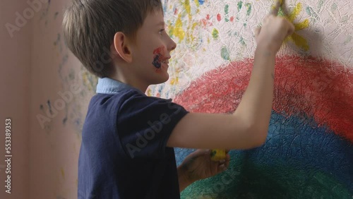 cheerful caucasian boy of 8-9 years old draws with paints with his hands on the wall of the house, selective focus. a child with a face stained with paints draws on the wall, a creative boy
