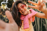 Bottom view of young caucasian woman sends peace and screeches hand at camera outdoor. Brunette wears sunglasses, sweater, top and jeans. Concept of funny moments, great vacation.