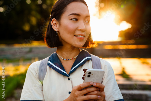 Joyful asian woman using smartphone while walking outdoors
