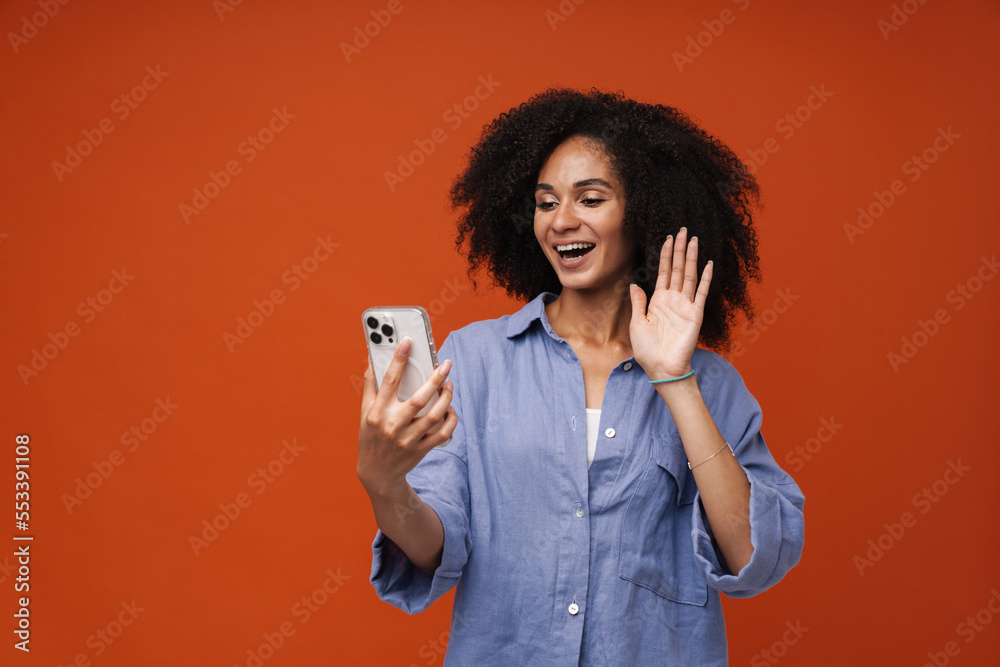 Cheerful afro woman talking by video call on smartphone and waving while standing isolated over red wall Cheerful afro woman talking by video call on smartphone and waving while standing isolated over red wall