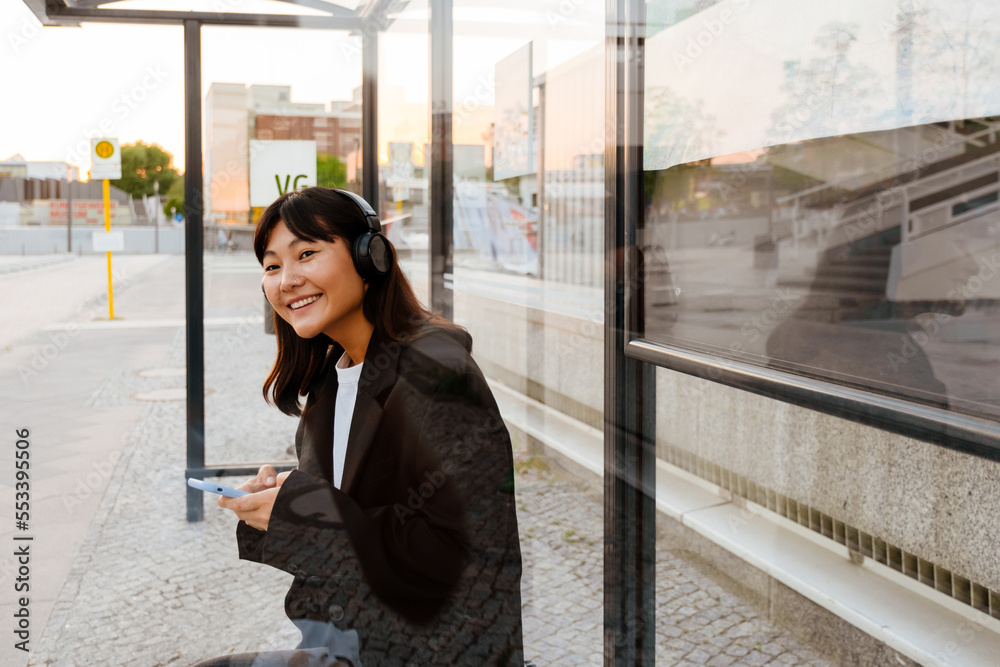 Fototapeta premium Young asian woman using cellphone and headphones sitting on bus station