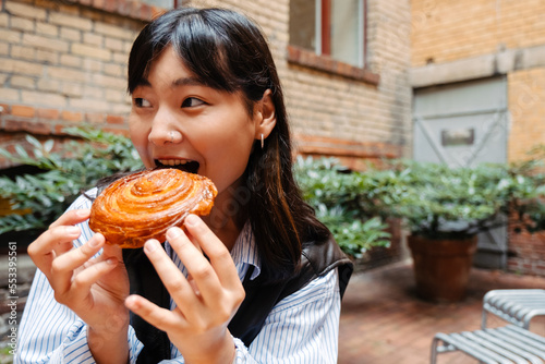 Young asian girl eating cinnabon bun while sitting in cafe