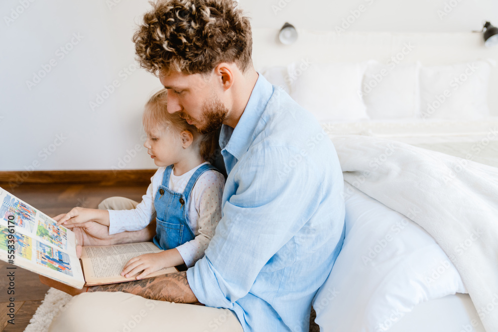 Young white father and little daughter reading book together at home