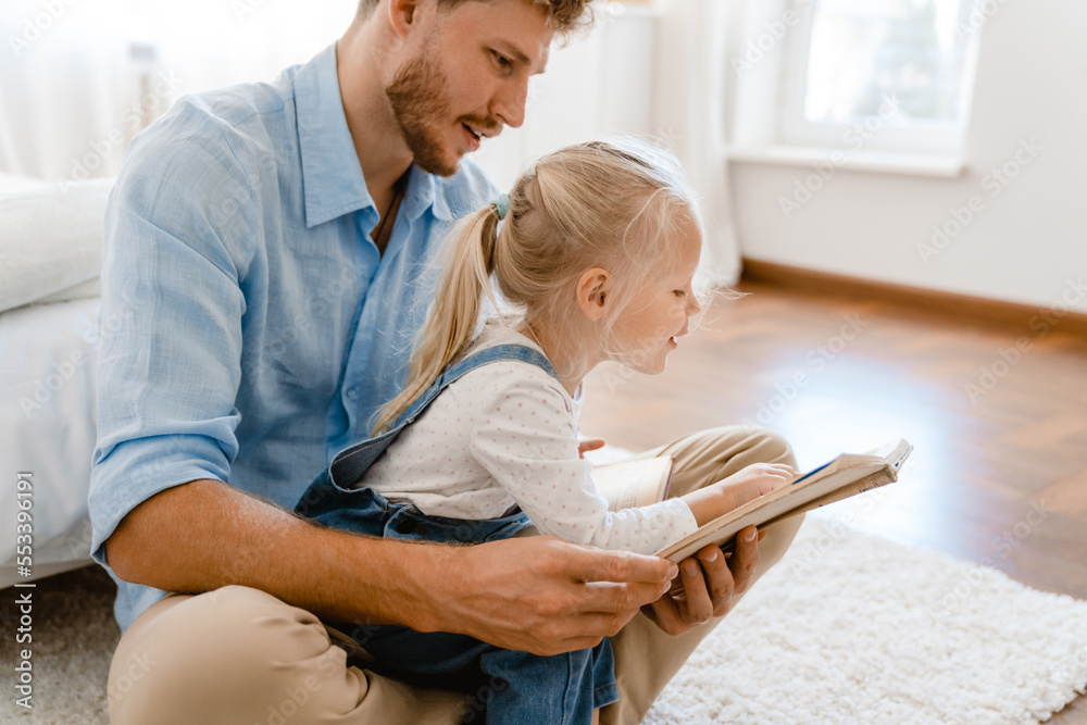 Fototapeta premium Young white father and little daughter reading book together at home