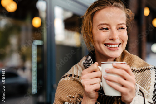 Joyful blonde woman in warm plaid drinking hot tea while stanging outdoors photo