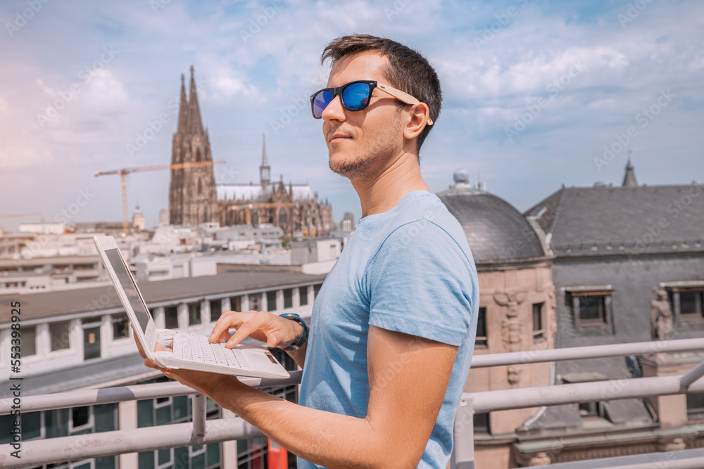 A young man remotely works on a laptop on an observation deck against the background of a view of Cologne Cathedral in Germany. Freelancer and student online concept A young man remotely works on a laptop on an observation deck against the background of a view of Cologne Cathedral in Germany. Freelancer and student online concept