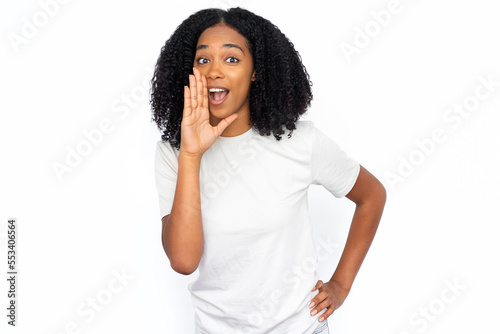 Excited African American woman shouting. Portrait of happy young female model with dark curly hair in white T-shirt looking at camera with hand near open mouth, calling someone. Advertisement concept